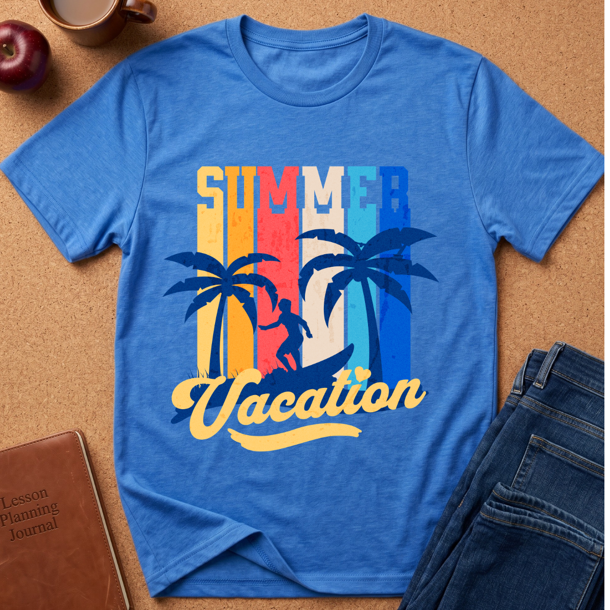 A blue short-sleeve graphic T-shirt laid flat on a cork background, featuring the words “SUMMER Vacation” with palm tree silhouettes and a retro striped background. Styled flat-lay props include a brown journal, a cup, an apple, and blue jeans.