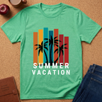 A mint green heathered t-shirt laid flat on a cork background, featuring a retro graphic of black palm trees in front of vertical orange, yellow, teal, and red stripes, with the text 'SUMMER VACATION' in white. Styled flat-lay props include an apple, a coffee cup, a brown journal, and blue jeans.
