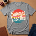 A heather gray short-sleeve t-shirt with a retro sunset graphic reading 'Summer Vacation' and 'EST 1998', laid flat on a cork background with an apple, coffee cup, notebook, and jeans nearby.