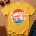 A yellow short-sleeve graphic T-shirt laid flat on a tan surface, with a retro sunset-style print reading “Summer Vacation” and “EST 1998.” Surrounding props include part of a red apple, a coffee cup, a brown notebook, and blue jeans.