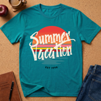 A teal short-sleeve graphic T-shirt laid flat on a cork surface, featuring a retro sunset design with the words 'Summer Vacation' in white script and 'EST 1998' below. Surrounding props include an apple, a coffee cup, a notebook, and folded blue jeans.