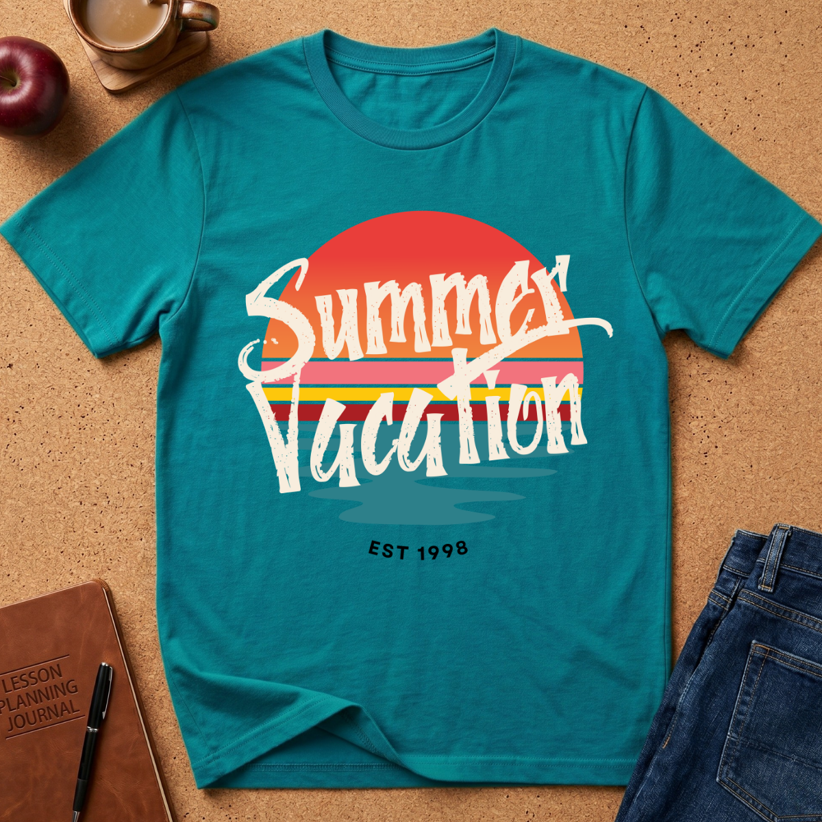 A teal short-sleeve graphic T-shirt laid flat on a cork surface, featuring a retro sunset design with the words 'Summer Vacation' in white script and 'EST 1998' below. Surrounding props include an apple, a coffee cup, a notebook, and folded blue jeans.