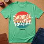 A green short-sleeve graphic T-shirt laid flat on a cork surface, with a retro sunset-style print reading “Summer Vacation” and “EST 1998.” Styled flat-lay props include an apple, a mug, a notebook, and blue jeans around the shirt.