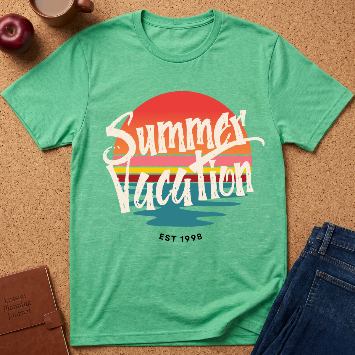 A green short-sleeve graphic T-shirt laid flat on a cork surface, with a retro sunset-style print reading “Summer Vacation” and “EST 1998.” Styled flat-lay props include an apple, a mug, a notebook, and blue jeans around the shirt.