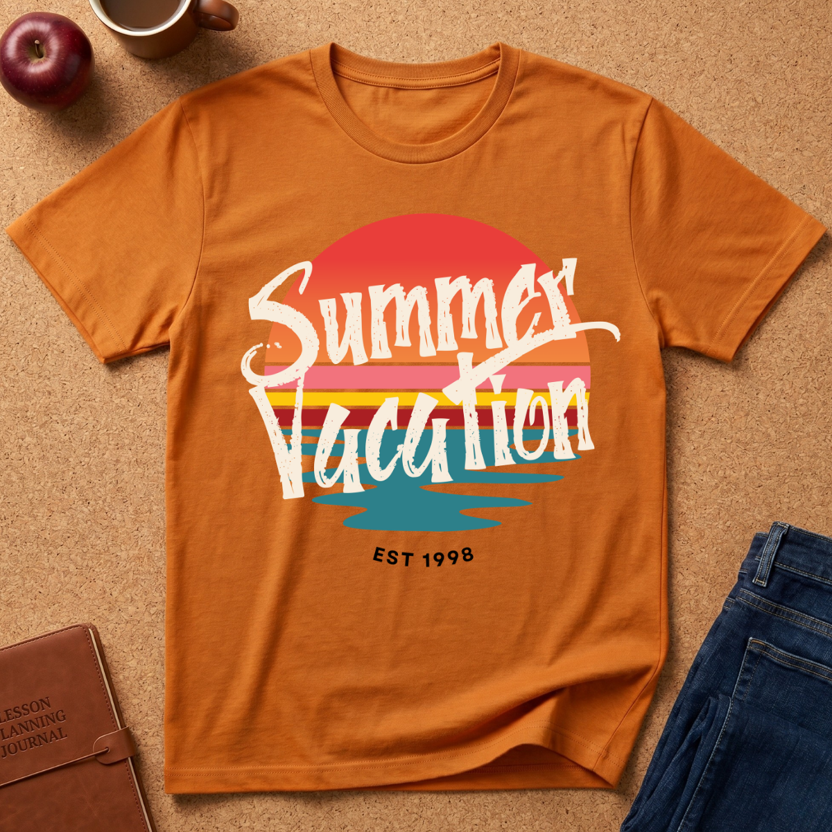 An orange graphic t-shirt laid flat on a cork background, featuring a retro sunset design with the words 'Summer Vacation' and 'EST 1998'. The shirt is styled with a brown leather journal, a red apple, a coffee mug, and blue jeans.