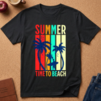 A black short-sleeve graphic T-shirt laid flat on a cork surface, featuring the words “SUMMER” and “TIME TO BEACH” in multicolor retro lettering above and below blue palm tree and surfer silhouettes. Styled flat-lay props include an apple, mug, journal, and jeans around the shirt.