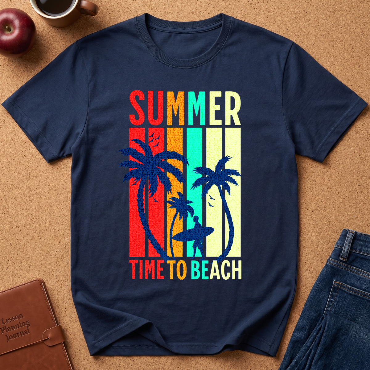 A navy blue short-sleeve t-shirt with a retro-style 'SUMMER TIME TO BEACH' graphic featuring palm trees and a surfer silhouette, laid flat on a brown cork background with styling props including an apple, coffee cup, and jeans.