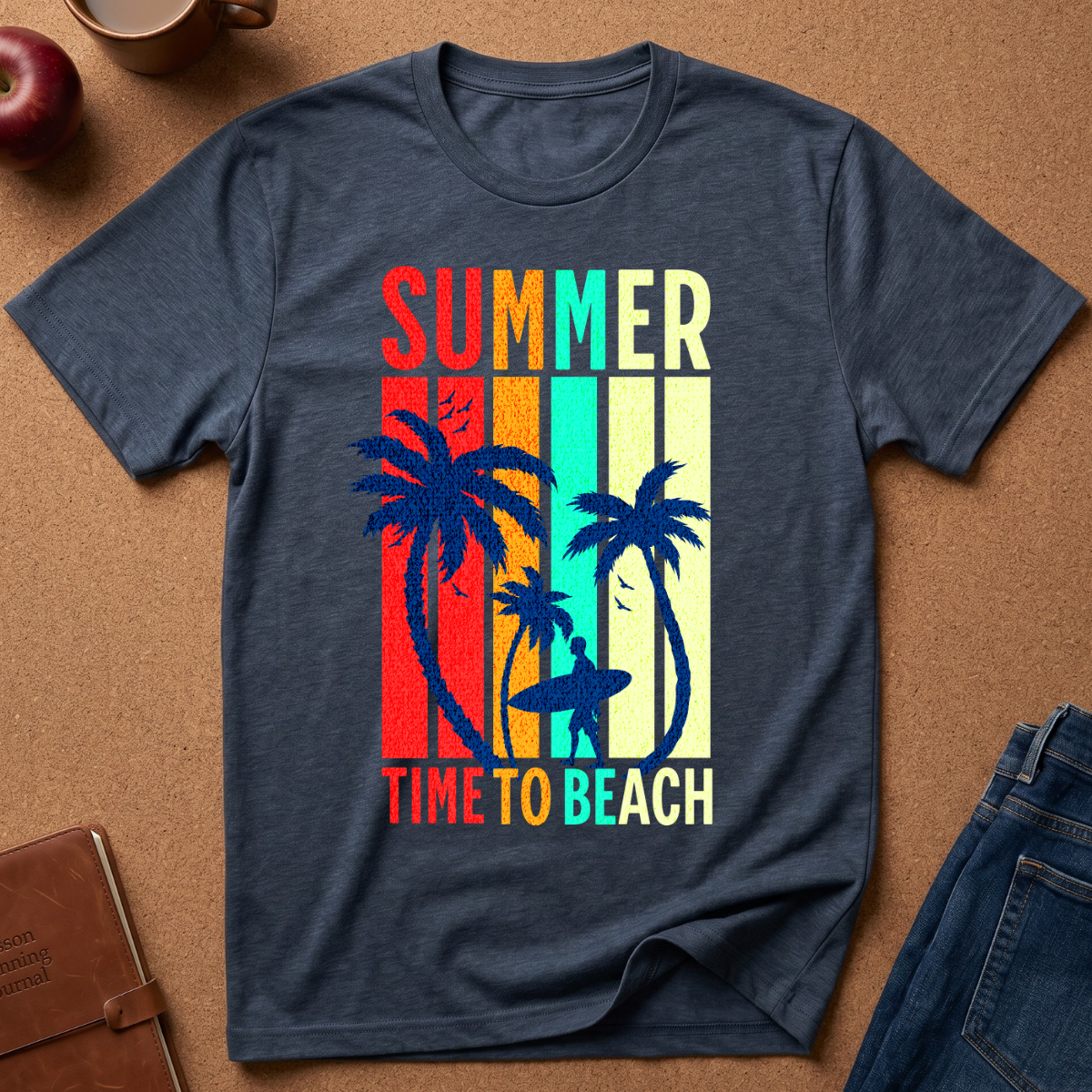 A heather navy blue t-shirt with a retro vertical-striped graphic featuring palm trees, surfers, and the text 'SUMMER TIME TO BEACH' in multicolor, laid flat on a cork background with jeans and an apple nearby.