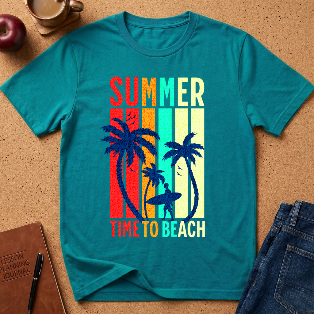 A teal short-sleeve graphic T-shirt laid flat on a cork surface, featuring the words 'SUMMER' and 'TIME TO BEACH' with palm trees and a surfer silhouette over vertical rainbow stripes. A red apple, coffee cup, notebook, and blue jeans are placed around it as styling props.