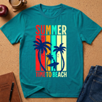 A teal short-sleeve graphic T-shirt laid flat on a cork surface, featuring the words 'SUMMER' and 'TIME TO BEACH' with palm trees and a surfer silhouette over vertical rainbow stripes. A red apple, coffee cup, notebook, and blue jeans are placed around it as styling props.