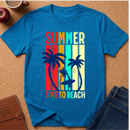 A bright blue short-sleeve t-shirt laid flat on a cork surface, featuring a retro vertical-striped graphic with palm trees and the words 'SUMMER TIME TO BEACH' in multicolor lettering. Surrounding props include an apple, a coffee cup, a brown journal, and folded blue jeans.