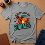 A heather gray short-sleeve t-shirt with a colorful retro graphic that reads 'SCHOOL'S OUT FOR SUMMER' with palm trees and a sunset, laid flat on a cork background with props including an apple, coffee cup, notebook, and jeans.