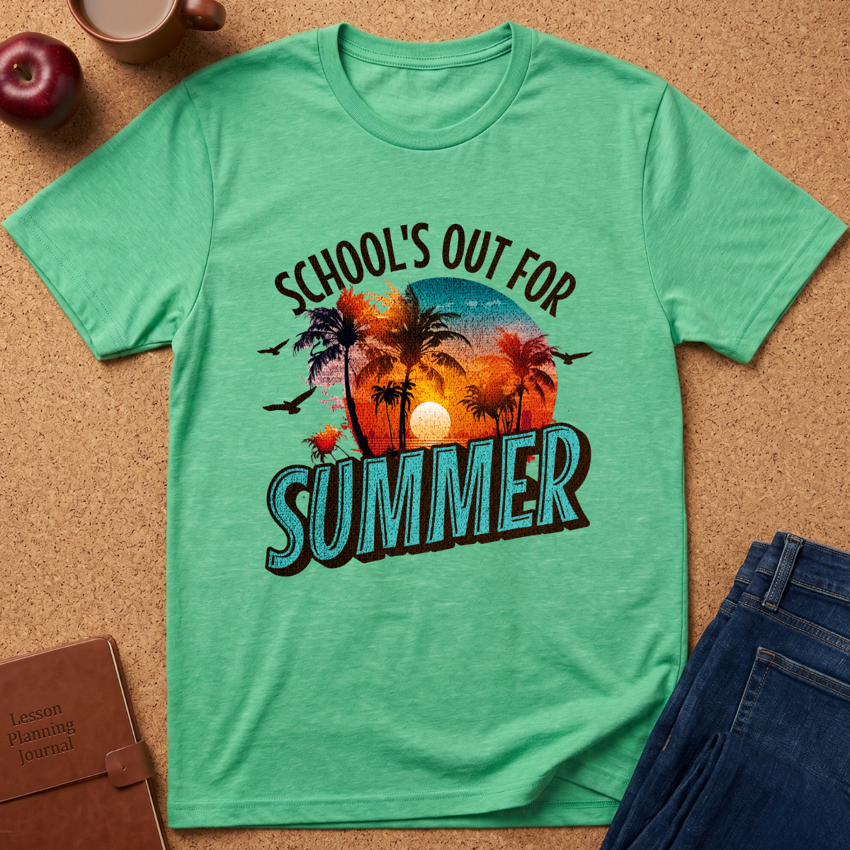 A mint green graphic t-shirt laid flat on a cork board background, styled with an apple, coffee cup, notebook, and jeans. The t-shirt features a colorful retro sunset graphic with palm trees and the text 'SCHOOL'S OUT FOR SUMMER'.