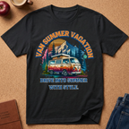 A black short-sleeve graphic T-shirt laid flat on a cork surface, featuring a colorful retro campsite scene with a van, trees, mountains, and the text 'VAN SUMMER VACATION' and 'DRIVE INTO SUMMER WITH STYLE.' Styled props include an apple, mug, journal, and jeans around the shirt.