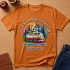 An orange short-sleeve t-shirt laid flat on a cork surface, featuring a colorful retro graphic of a van in front of mountains and trees with the text 'VAN SUMMER VACATION' and 'DRIVE INTO SUMMER WITH STYLE.' Props around it include an apple, a mug, a notebook, and blue jeans.