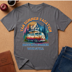 A gray short-sleeve graphic T-shirt laid flat on a cork surface, surrounded by lifestyle props including an apple, coffee cup, brown journal, and blue jeans. The shirt features a colorful retro-style mountain and campervan design with the text 'VAN SUMMER VACATION' and 'DRIVE INTO SUMMER WITH STYLE.