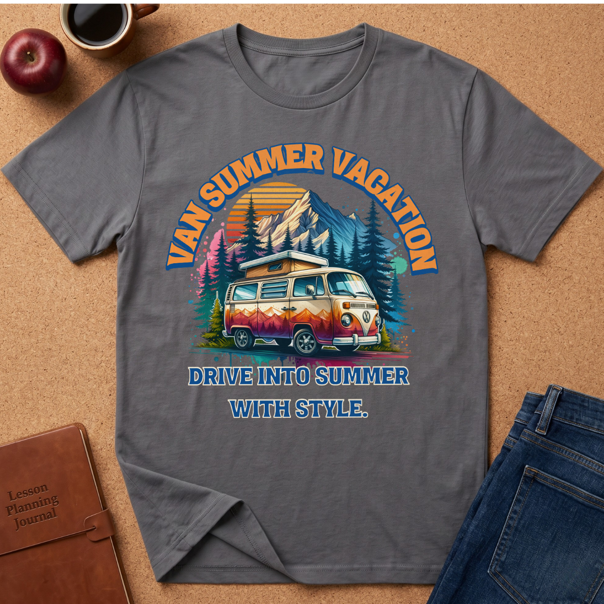 A gray short-sleeve graphic T-shirt laid flat on a cork surface, surrounded by lifestyle props including an apple, coffee cup, brown journal, and blue jeans. The shirt features a colorful retro-style mountain and campervan design with the text 'VAN SUMMER VACATION' and 'DRIVE INTO SUMMER WITH STYLE.