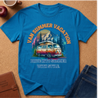 A bright blue graphic t-shirt laid flat on a cork background, featuring a retro-style illustration of a vintage van with mountains, trees, and the text 'VAN SUMMER VACATION' and 'DRIVE INTO SUMMER WITH STYLE.' Surrounding props include an apple, a coffee cup, a brown journal, and folded blue jeans.