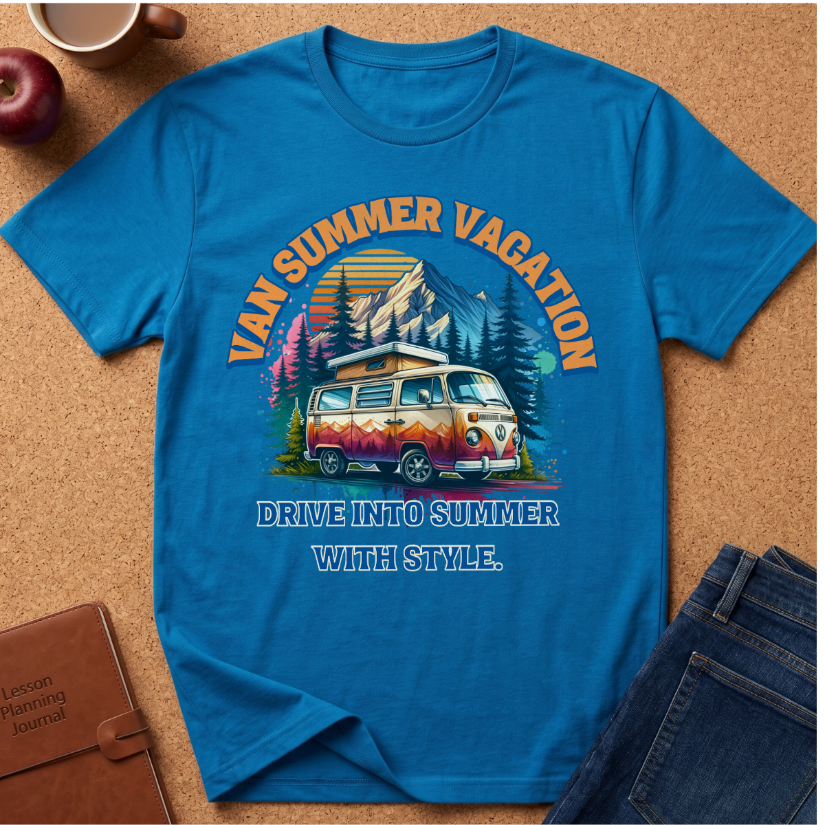 A bright blue graphic t-shirt laid flat on a cork background, featuring a retro-style illustration of a vintage van with mountains, trees, and the text 'VAN SUMMER VACATION' and 'DRIVE INTO SUMMER WITH STYLE.' Surrounding props include an apple, a coffee cup, a brown journal, and folded blue jeans.