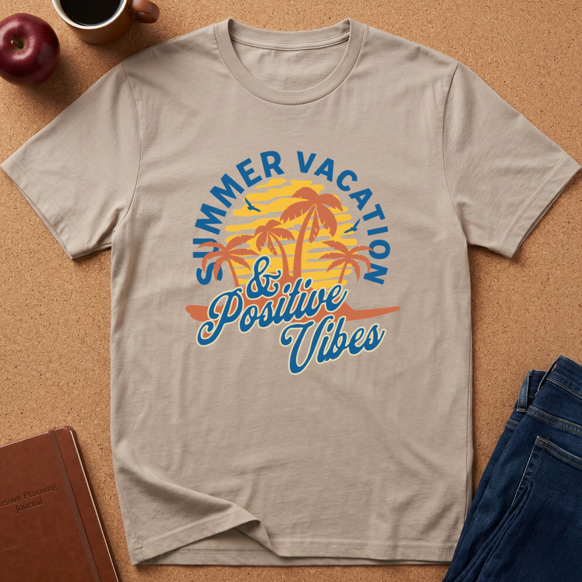 A light beige crewneck t-shirt with a colorful retro graphic reading 'SUMMER VACATION & Positive Vibes' around palm trees, styled in a flat lay on a cork background with a coffee mug, apple, notebook, and jeans.