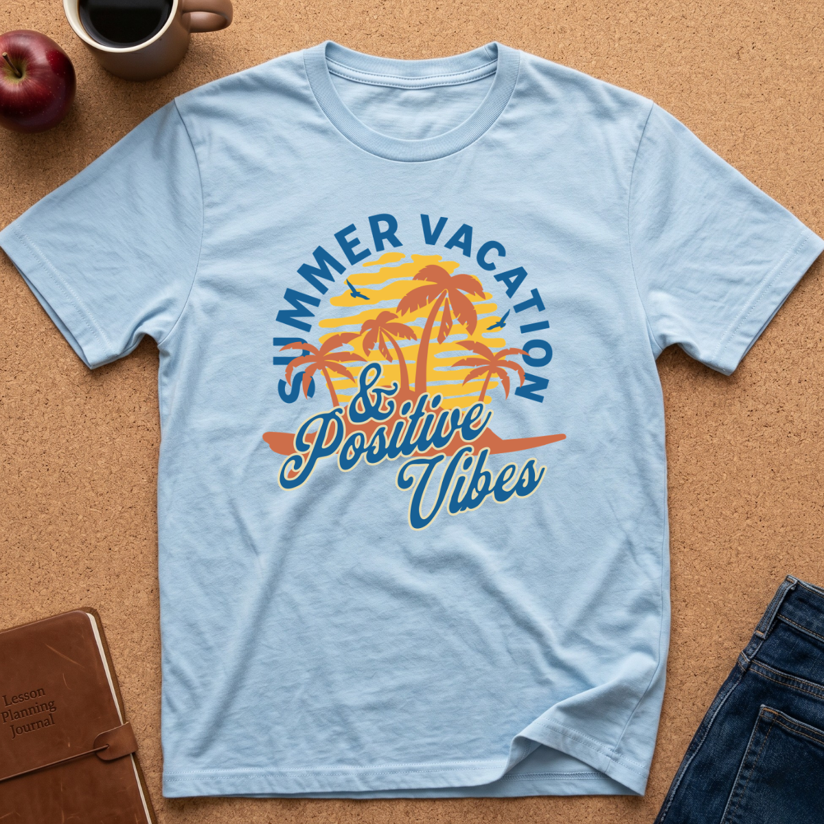 A light blue short-sleeve t-shirt laid flat on a cork background, featuring a colorful graphic of palm trees, a yellow sun, and the text 'SUMMER VACATION & Positive Vibes' in blue and orange. Styled flat-lay props include a coffee mug, an apple, a notebook, and blue jeans.
