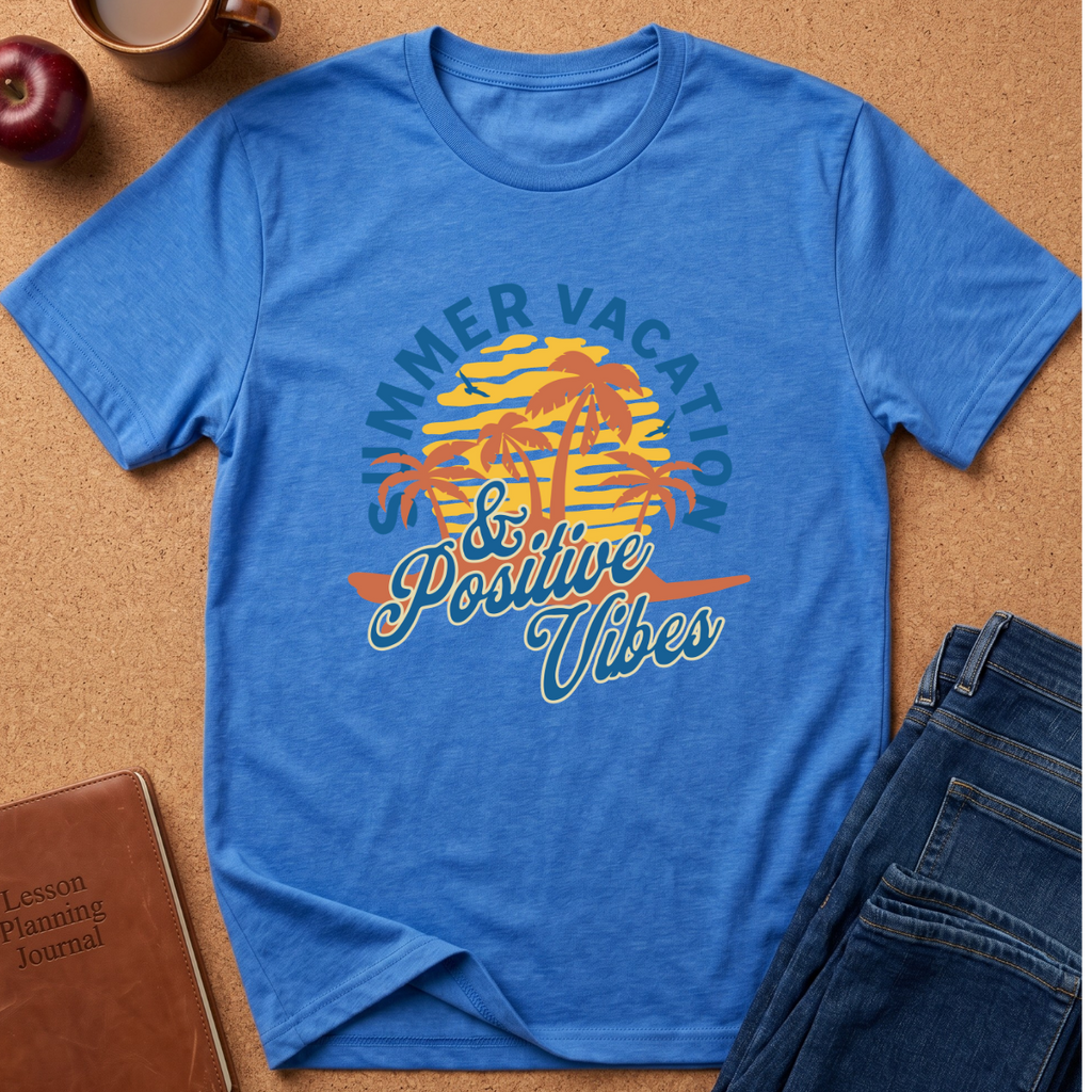 A blue short-sleeve graphic T-shirt laid flat on a cork surface, surrounded by styled props including an apple, a coffee mug, a brown journal, and folded blue jeans. The shirt features a retro beach sunset graphic with palm trees and the text 'Summer Vacation & Positive Vibes' in blue, yellow, and orange.
