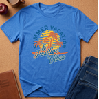 A blue short-sleeve graphic T-shirt laid flat on a cork surface, surrounded by styled props including an apple, a coffee mug, a brown journal, and folded blue jeans. The shirt features a retro beach sunset graphic with palm trees and the text 'Summer Vacation & Positive Vibes' in blue, yellow, and orange.
