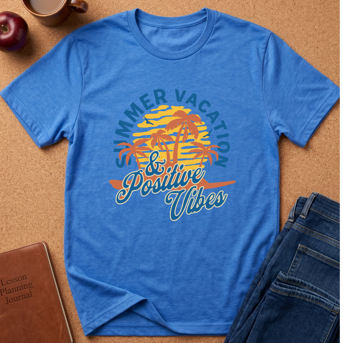 A blue short-sleeve graphic T-shirt laid flat on a cork surface, surrounded by styled props including an apple, a coffee mug, a brown journal, and folded blue jeans. The shirt features a retro beach sunset graphic with palm trees and the text 'Summer Vacation & Positive Vibes' in blue, yellow, and orange.