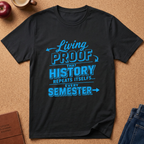 A black short-sleeve graphic T-shirt laid flat on a cork surface, with blue text reading 'Living PROOF THAT HISTORY REPEATS ITSELF.