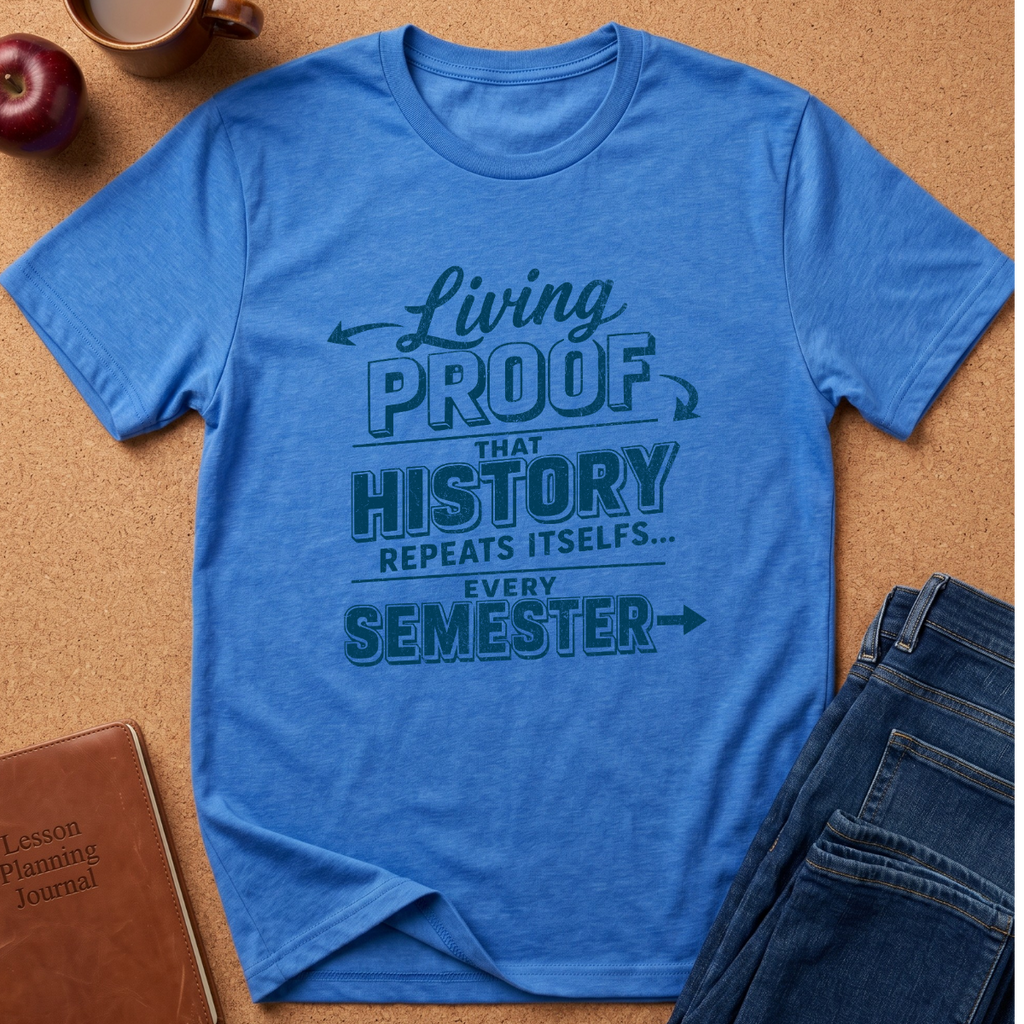 A blue graphic t-shirt laid flat on a cork background, styled with a brown leather journal, a red apple, and folded blue jeans. The t-shirt features a dark teal graphic text that reads 'Living PROOF THAT HISTORY REPEATS ITSELF.