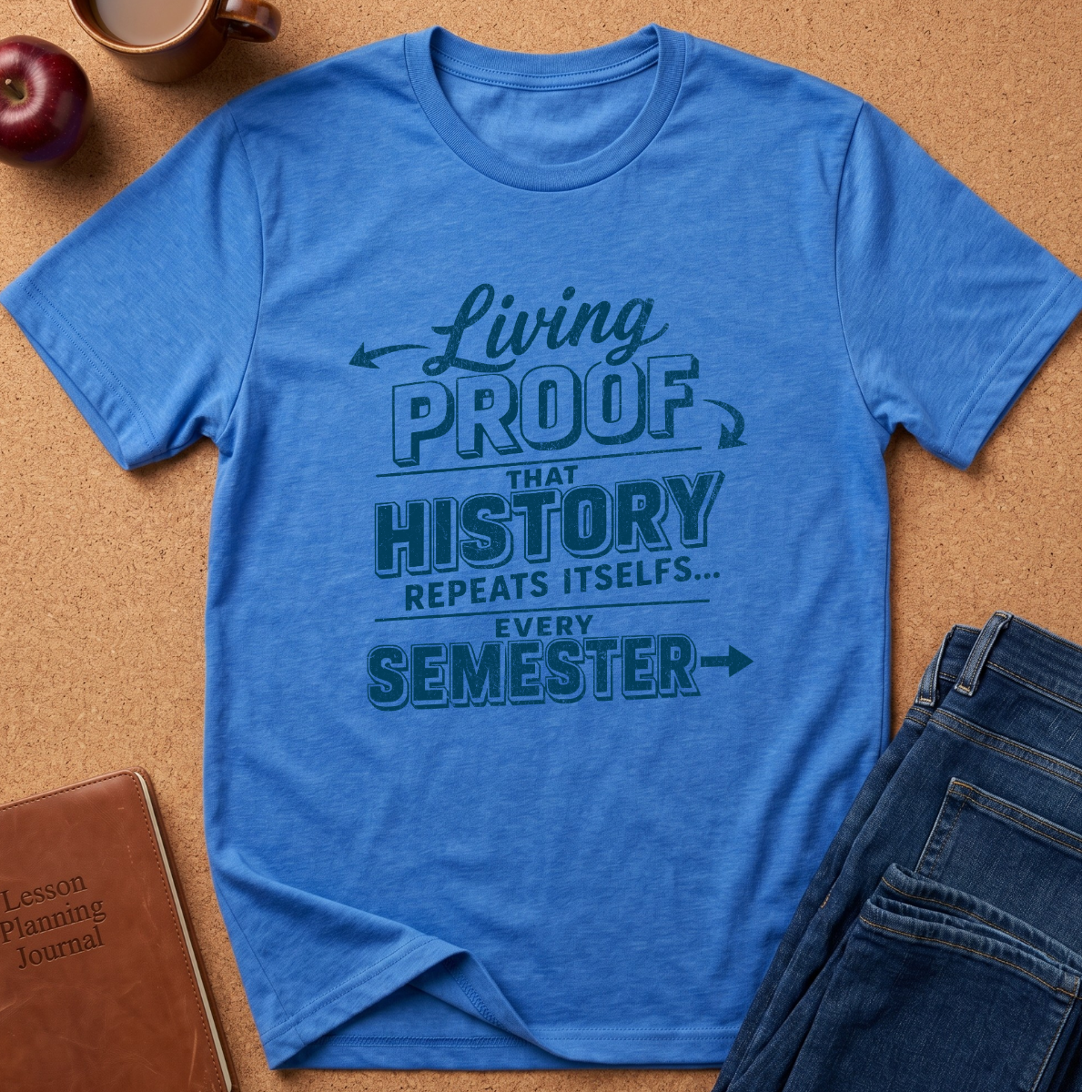 A blue graphic t-shirt laid flat on a cork background, styled with a brown leather journal, a red apple, and folded blue jeans. The t-shirt features a dark teal graphic text that reads 'Living PROOF THAT HISTORY REPEATS ITSELF.
