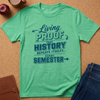 A mint green graphic t-shirt laid flat on a cork background, surrounded by lifestyle props including an apple, a coffee mug, a brown journal, and blue jeans. The t-shirt features a dark teal graphic text that reads 'Living PROOF THAT HISTORY REPEATS ITSELF.