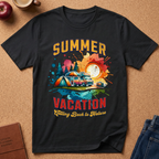 A black short-sleeve graphic T-shirt laid flat on a cork-like surface, featuring a colorful summer vacation camping design with a tent, camper van, trees, and the text 'SUMMER VACATION Calling Back to Nature.' Partial props around it include an apple, a mug, a journal, and jeans.