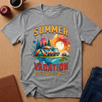 A heather gray graphic t-shirt laid flat on a cork background, featuring a colorful retro-style illustration with the words 'SUMMER VACATION' and a camping scene. Styled flat-lay props include a coffee cup, apple, notebook, and jeans.