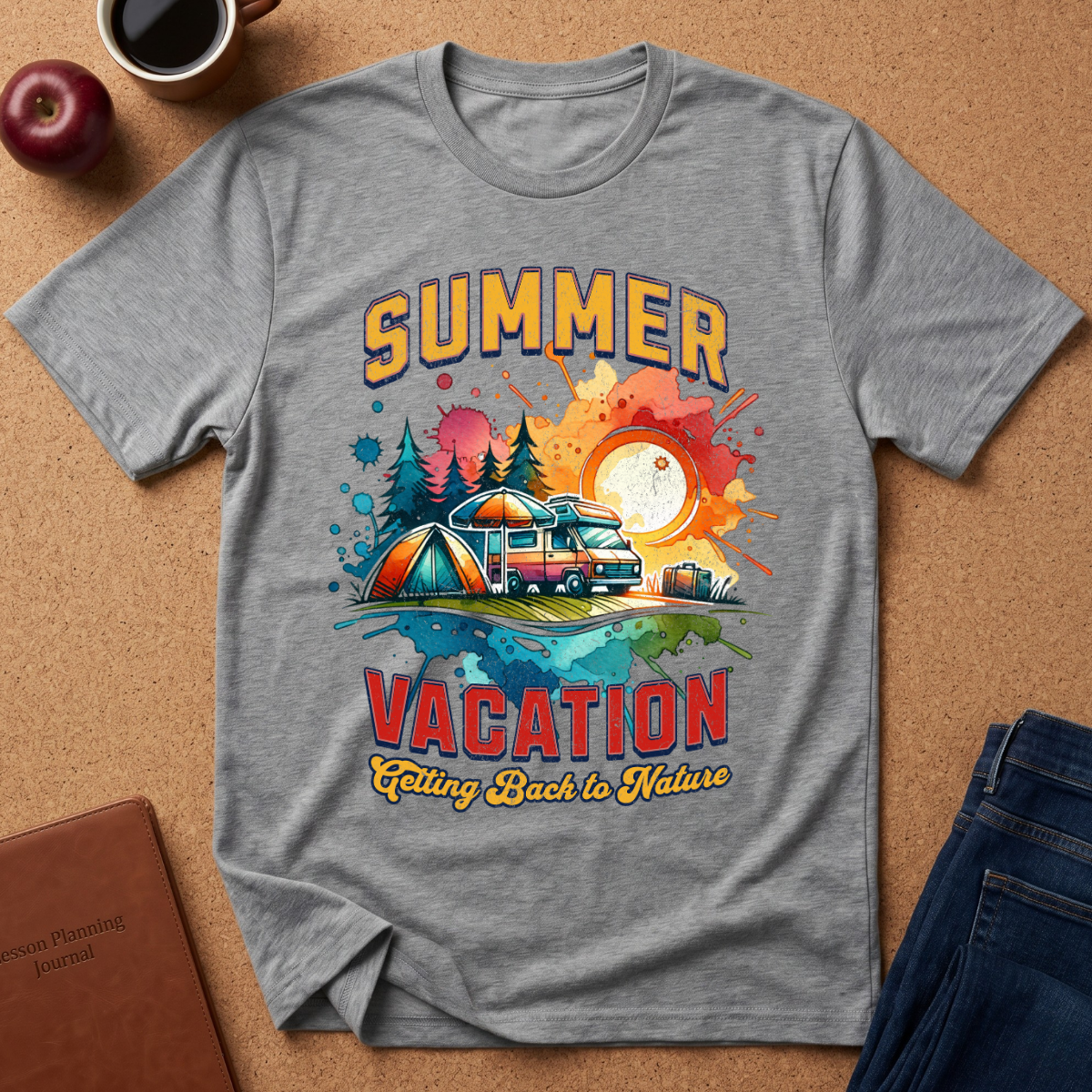 A heather gray graphic t-shirt laid flat on a cork background, featuring a colorful retro-style illustration with the words 'SUMMER VACATION' and a camping scene. Styled flat-lay props include a coffee cup, apple, notebook, and jeans.