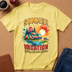 A yellow short-sleeve graphic T-shirt laid flat on a cork-like surface, surrounded by a coffee cup, apple, notebook, and folded jeans. The shirt features a colorful retro-style travel graphic with the words “SUMMER VACATION” and an illustration of a camper van, tent, trees, and sun.
