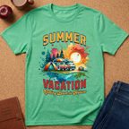 A mint green short-sleeve t-shirt laid flat on a cork background, featuring a colorful retro-style graphic with the words 'SUMMER VACATION' and 'Falling Back to Nature' around an illustration of a campsite scene with a tent, camper, trees, and sunset. Partial jeans, a book, and a cup are visible around the shirt as styling props.