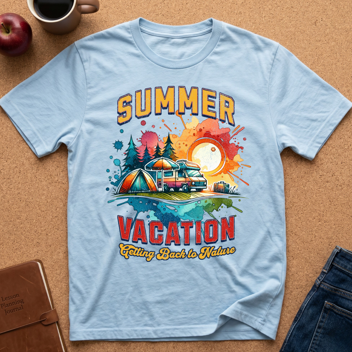 A light blue short-sleeve t-shirt laid flat on a cork background, featuring a colorful graphic print with the words 'SUMMER VACATION' and an illustration of a campsite scene with a tent, camper van, trees, and sunset. A coffee mug, apple, notebook, and jeans are partially visible around it as styling props.