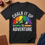 A black short-sleeve graphic T-shirt laid flat on a cork surface, featuring the phrase “CHALK IT UP TO ADVENTURE” in white text above and below a multicolor mountain-and-pencil design. Partial props around the shirt include an apple, a mug, a journal, and jeans.