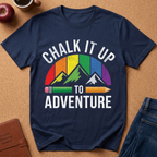 A navy blue graphic t-shirt laid flat on a cork background, featuring a colorful mountain design with a rainbow arch and the words 'CHALK IT UP TO ADVENTURE' in white. Styled flat-lay props include an apple, a coffee cup, a brown journal, and blue jeans.