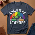 A heather navy blue graphic t-shirt laid flat on a brown cork background. The shirt features a colorful retro design with the text 'CHALK IT UP TO ADVENTURE' and a mountain graphic filled with rainbow colors and a pencil motif.