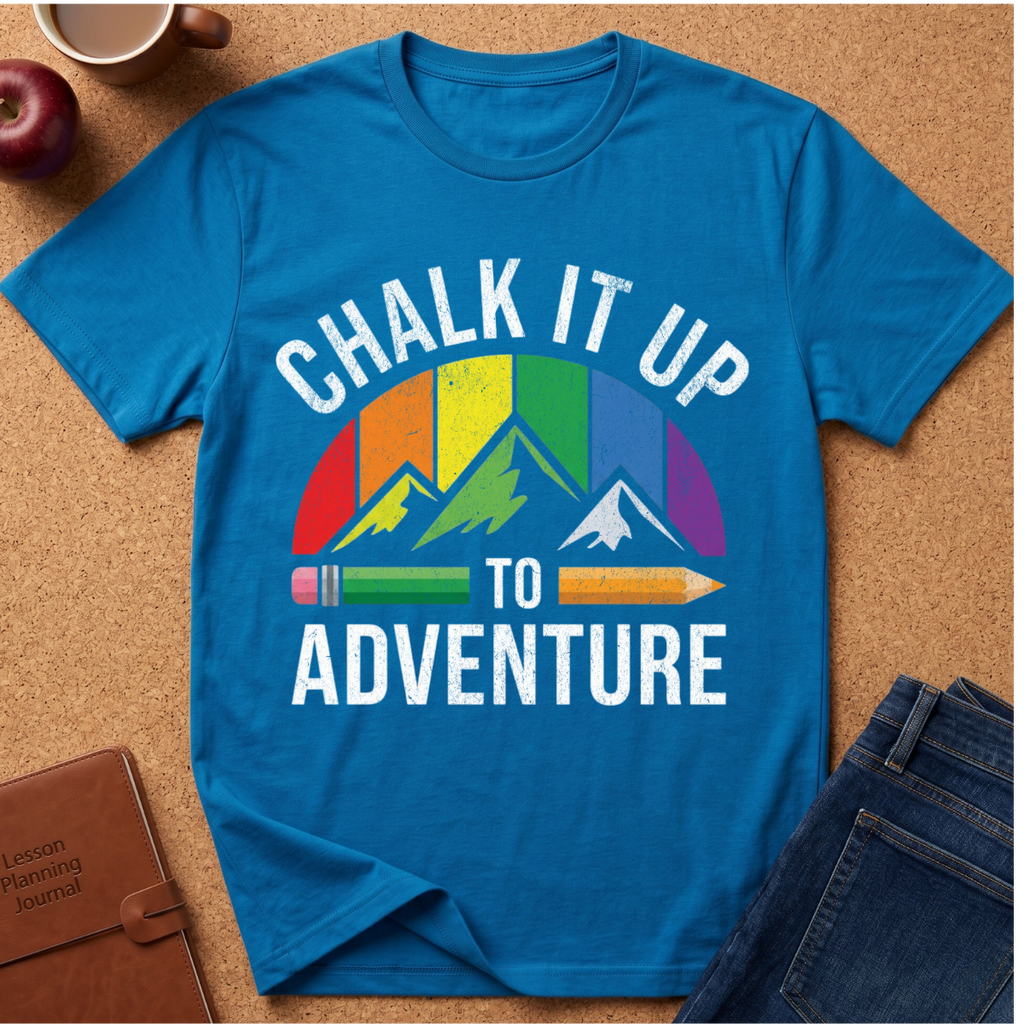 A bright blue graphic T-shirt laid flat on a cork surface, surrounded by styled props including an apple, a mug, a journal, and jeans. The shirt features the phrase “CHALK IT UP TO ADVENTURE” in white text above and below a mountain graphic with rainbow-colored stripes and pencils.