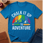 A bright blue graphic T-shirt laid flat on a cork surface, surrounded by styled props including an apple, a mug, a journal, and jeans. The shirt features the phrase “CHALK IT UP TO ADVENTURE” in white text above and below a mountain graphic with rainbow-colored stripes and pencils.