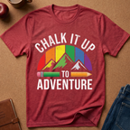 A heathered red short-sleeve t-shirt laid flat on a brown textured surface, featuring a colorful retro mountain graphic with the words 'CHALK IT UP TO ADVENTURE' in white. Styled flat-lay props include a brown notebook, a blue denim jacket, an apple, and a mug.