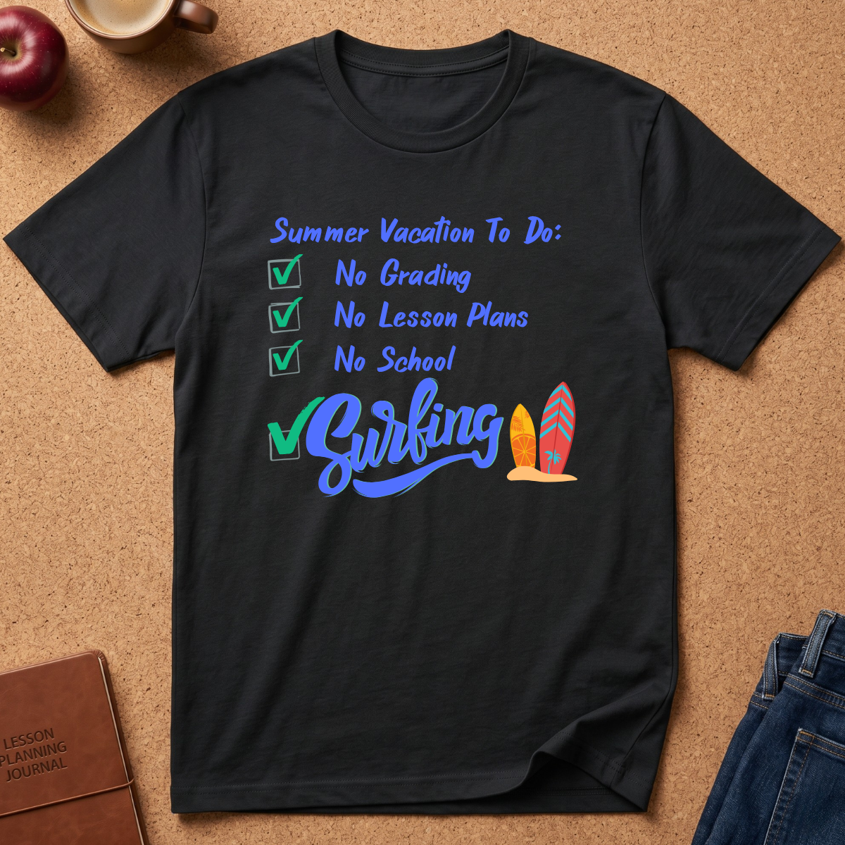 A black short-sleeve graphic T-shirt laid flat on a cork-like surface, with a vacation checklist design in blue and green reading 'Summer Vacation To Do: No Grading, No Lesson Plans, No School' and 'Surfing' with two colorful surfboards. Partial props around the shirt include an apple, a mug, a journal, and jeans.