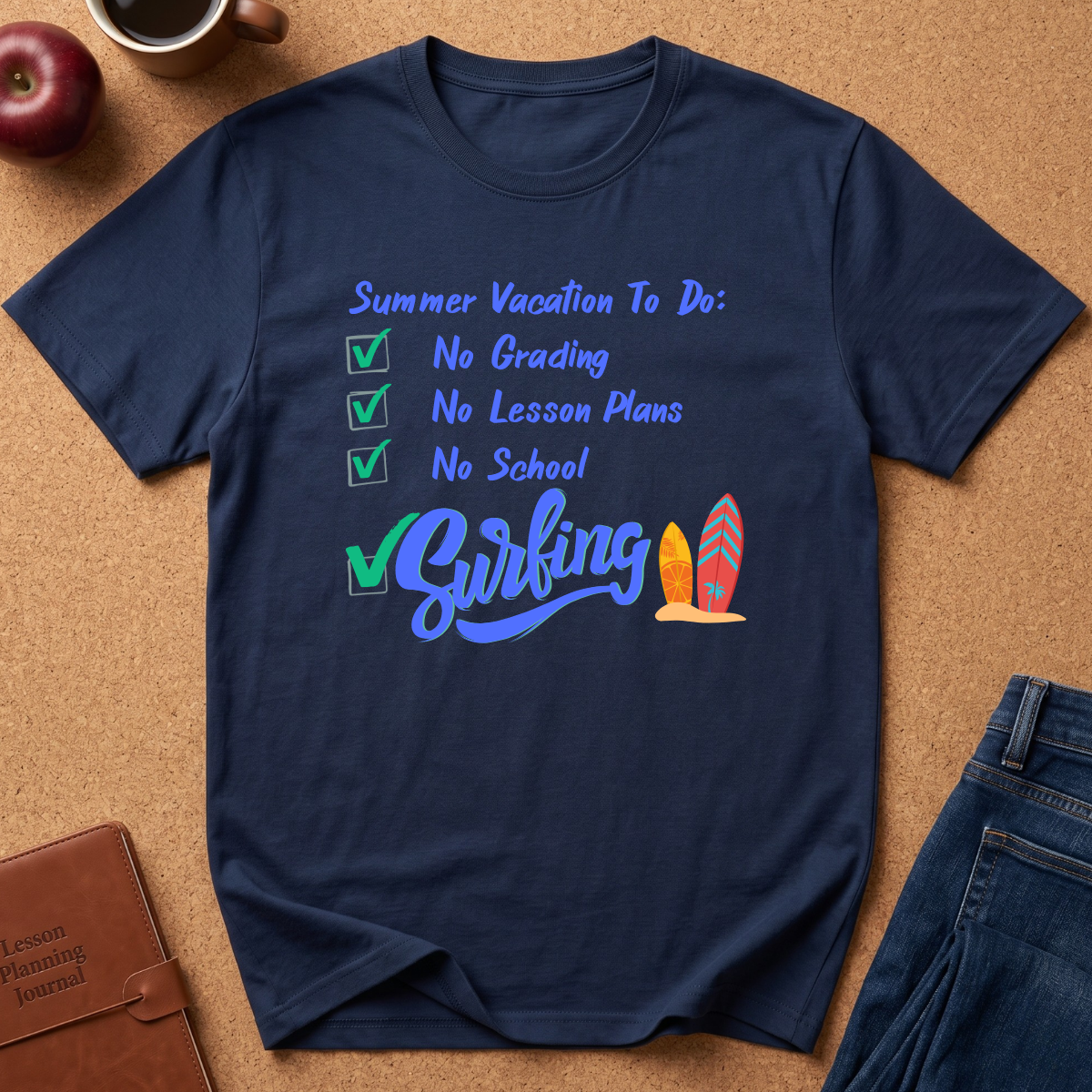 A navy blue graphic t-shirt laid flat on a cork board background, featuring a checklist design for summer vacation with 'No Grading', 'No Lesson Plans', 'No School', and 'Surfing' in blue script alongside two colorful surfboards. Partial jeans, an apple, a coffee cup, and a journal are visible around the shirt as styled props.