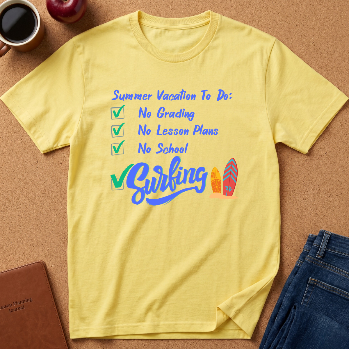 A yellow short-sleeve t-shirt laid flat on a cork background, featuring a blue and green graphic that reads 'Summer Vacation To Do: No Grading, No Lesson Plans, No School' and 'Biking' with two surfboards. A coffee mug, apple, notebook, and jeans are styled around it.