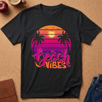 A black short-sleeve t-shirt laid flat on a cork background, featuring a retro sunset graphic with palm trees and the words 'beach vibes' in pink and orange lettering. Styled flat-lay props include an apple, a coffee cup, a journal, and jeans.