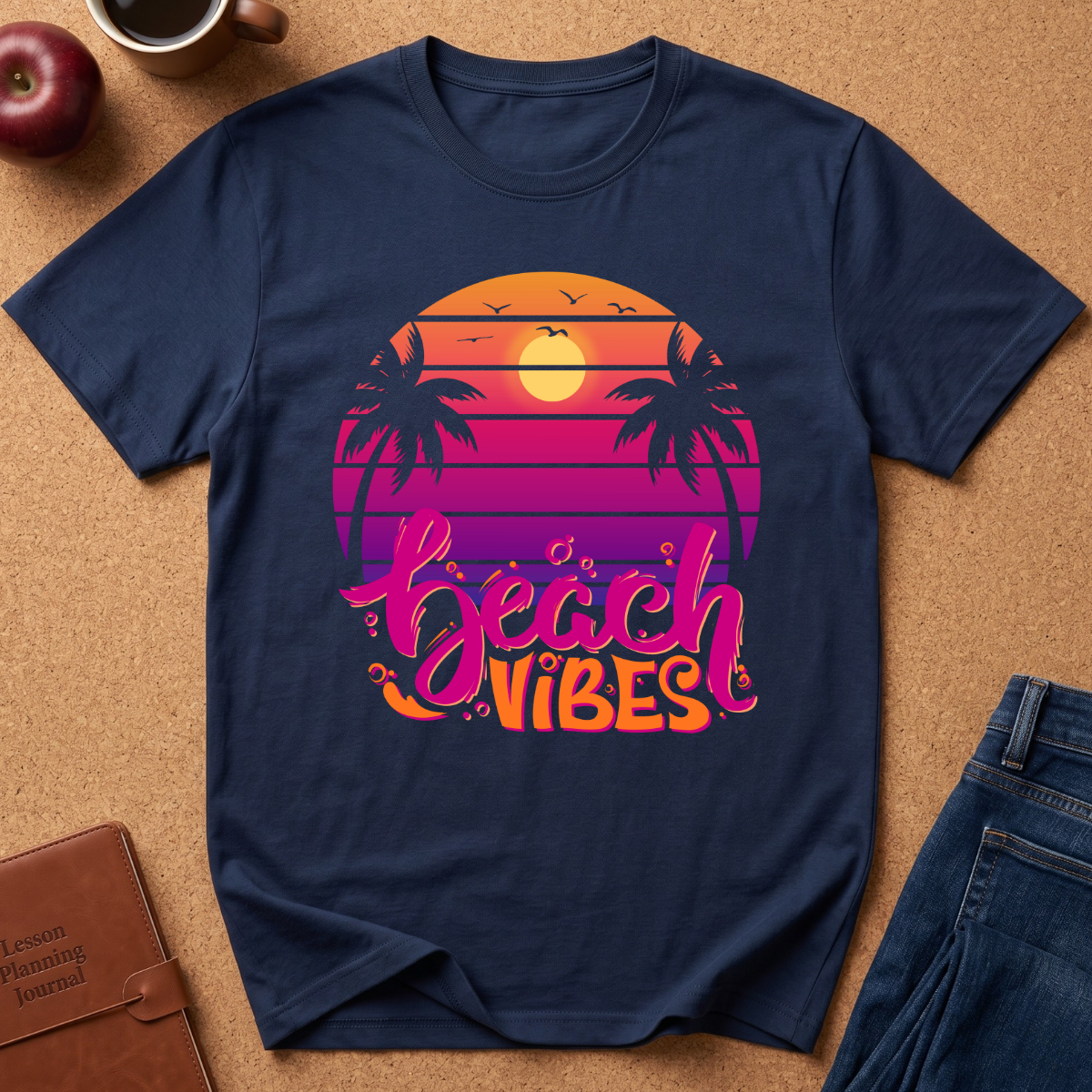 A navy blue short-sleeve t-shirt with a retro sunset graphic on the chest. The graphic features a circular sunset gradient in orange, yellow, pink, and purple with palm tree silhouettes and the text 'beach VIBES' in stylized pink and orange lettering.