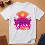 A white short-sleeve t-shirt laid flat on a cork surface, featuring a retro sunset graphic with palm trees and the words 'beach vibes' in pink and orange lettering. Styled flat-lay props include an apple, a coffee cup, a notebook, and jeans.